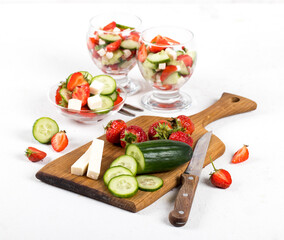 Ingredients, the process of preparing a berry and vegetable salad of strawberries and cucumber with the addition of feta cheese on a white background