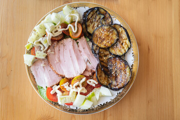 Salad on a round plate seen from above on a wooden table. The meal contains sliced ham, fried sliced aubergine, cucumber and tomato.