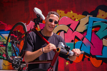 a guy with round sunglasses in a dark t-shirt and shorts with a bicycle against a bright wall with graffiti