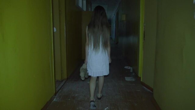 a girl with long black hair and a white dress is walking slowly in a dark corridor. The concept of a horror maniac. Halloween. she is holding a Teddy bear,