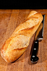 Top view, close distance of a freshly baked, french baguette  and a serrated edge knife with a black handle on a wood cutting board