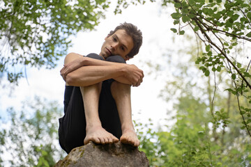Low angle portrait of man hugging knees while sitting on rock in forest