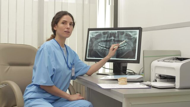 Medium shot of professional female dentist wearing medical overall sitting at desk, holding pen in her hand and examining x-ray image of jaw on computer screen