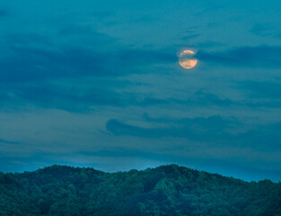 Full moon behind clouds in blue sky over mountains covered with lush green foliage. Taken at blue hour just before sunset.