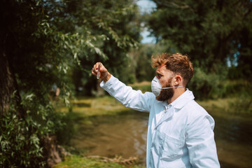 Male caucasian scientist biologist and researcher in protective suit with mask taking water samples from polluted river.