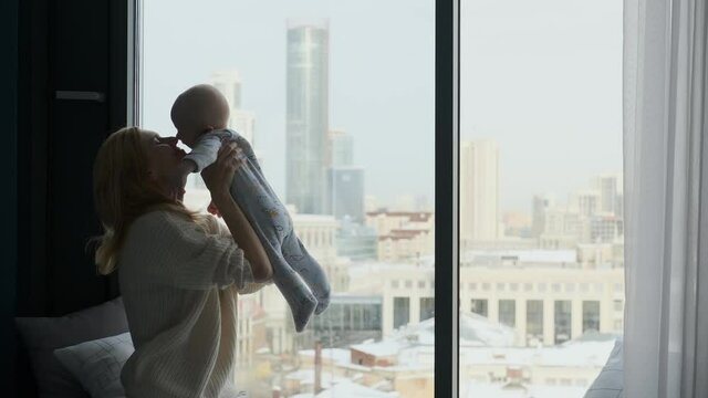 Happy Mom Picks Up And Hugs Baby Sitting By The Window