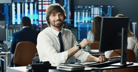 Portrait of male stock trader operating at hiis workstation using headset and computer on background of multiple monitors showing data, ticker numbers and graphs. Business Team, Trading Concept