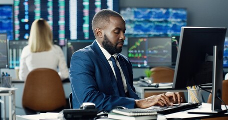 Portrait of african american male stock trader working at stock exchange office using computer on background of his business team and multiple monitors showing data, ticker numbers and graphs.