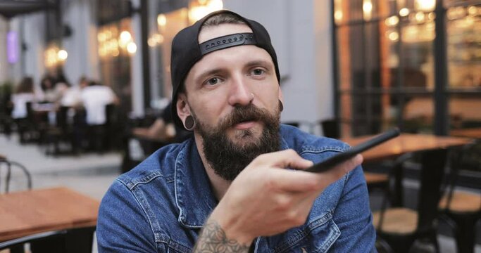 Caucasian handsome young male hipster with beard and piercing sitting outdoor at table on cafe terrace and recording voice message on smartphone. Guy talking and sending audio on mobile phone.