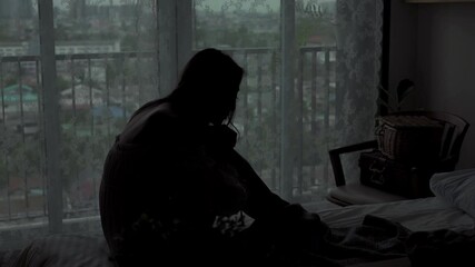 4K Silhouette of Depressed Young beautiful Asian woman sitting alone on the bed in apartment bedroom with crying. Loneliness sad girl think of grief trouble. Negative emotion and mental health concept