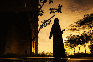 a young woman wearing a hijab and jilbab standing holding a phone near a water body at dusk