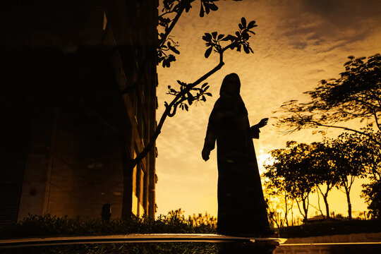 a young woman wearing a hijab and jilbab standing holding a phone near a water body at dusk