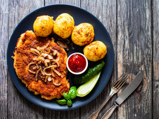 Breaded pork chop with boiled potatoes and cucumber served on black plate on wooden table