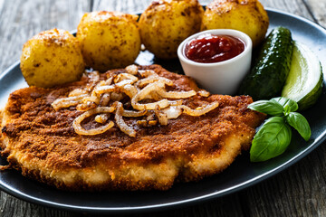 Breaded pork chop with boiled potatoes and cucumber served on black plate on wooden table