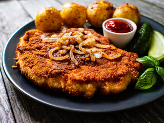 Breaded pork chop with boiled potatoes and cucumber served on black plate on wooden table