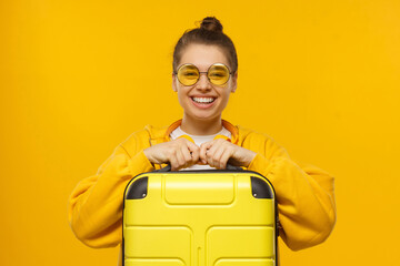 Young funny smiling girl in hoodie and colored glasses holding travel suitcase, willing to go on...