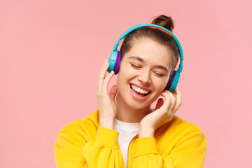 Young smiling teen girl dressed in yellow hoodie, enjoying favorite music through wireless headphones with closed eyes, isolated on pink background