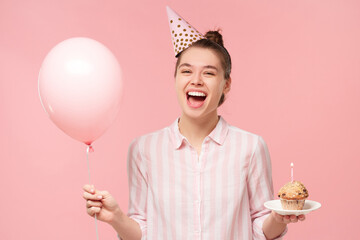 Young excited happy teen girl wearing birthday hat, holding balloon and plate with cake, isolated...