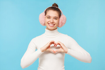 Teen girl in white sweater and pink earmuffs, showing heart gesture with hands, sharing love and care, isolated on blue background