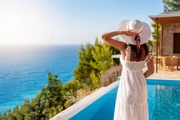 Eine elegante Frau mit Hut steht am Rande eines Pools und schaut auf das blaue Meer Griechenlands wöhrend ihres Sommerurlaubes