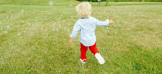 Unknown child cheerfully runs away on the green floor behind small soap bubbles