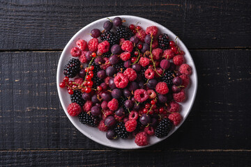 Tasty fresh ripe raspberry, blackberry, gooseberry and red currant berries in plate, healthy food fruit on dark wooden background, top view