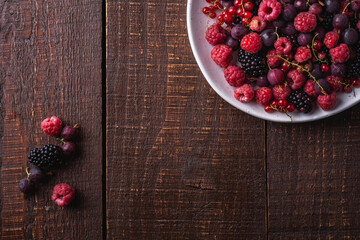 Tasty fresh ripe raspberry, blackberry, gooseberry and red currant berries in plate, healthy food fruit on dark brown wooden background, top view copy space