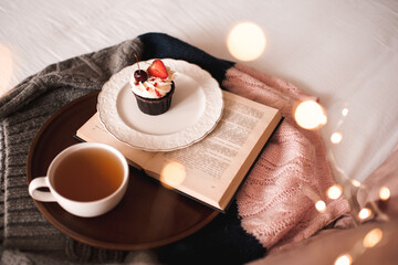 Cup of green tea with tasty cupcake and open book on knitted sweater in bed closeup. Good morning. Selective focus.