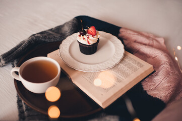 Cup of green tea with tasty cupcake and open book on knitted sweater in bed closeup. Good morning. Selective focus.
