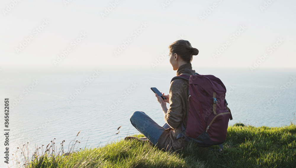 Wall mural Young tourist woman using smartphone over beautiful landscape with sea. Traveler girl with backpack and mobile phone. Travel, tourism, summer holidays, active lifestyle concept