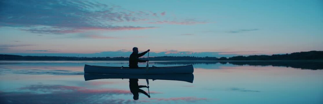 Man canoeing in a traditional wooden boat on a large lake at dawn. Shot on RED cinema camera with 2x anamorphic lens