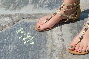 Naklejka premium Cadaqués, Spain, 11 July 2020.A view of a woman's feet in golden sandals reads a sentence about LGTBIQ Pride Day that has been written on a stone floor by the sea.