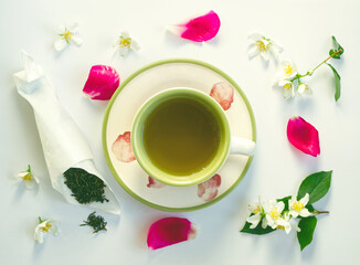 Teacup of green jasmine tea with jasmine flowers, leaves, bag of tea and pink petals on white background. Beautiful creme cup and tea plate with soft pink petals. 