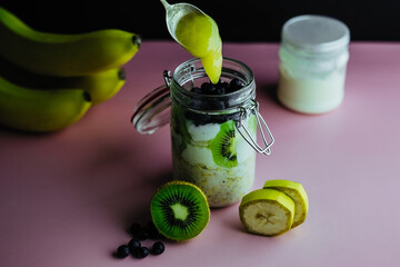 Overnight oatmeal in a jar. Healthy breakfast and diet. with kiwi, banana, blueberry and greek yogurt. On pink background. Honey on spoon.  Copy space.