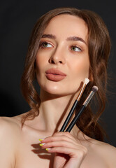 Portrait of the beautiful brunette woman with make-up brushes near face. Girl posing over Gray background. Clean natural makeup.