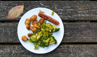 Vegan meal with oven baked potatos, plant based sausace, broccoli, peas and pesto on a white plate.