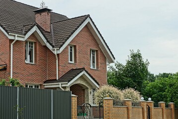 facade of a brown brick private house with windows under a tiled roof behind the fence