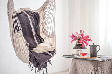 A boho-style hammock chair with a stack of books. Wooden tray with teapot and Cup of tea and beautiful Lily flowers. Cozy interior. The concept of rest and home comfort.