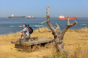 A sawn-off tree is focused at a viewing point on the Strait of Gibraltar. Behind it out of focus with bokeh a hiker and many large ships at sea. - obrazy, fototapety, plakaty