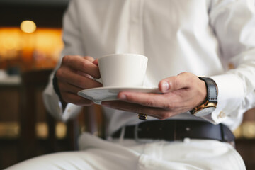 Fototapeta na wymiar An elegant man in a white suit drinks coffee from a white mug. Hands and mug close -up