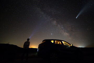 Silhouette of a man with head flashlight standing beside a car  lighting a beam of light on Milkyway galaxy and Neowise comet with light tail in dark night sky.