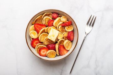 Trendy home breakfast with tiny cereal pancakes with piece of butter and fresh strawberries in the bowl