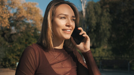 Beautiful smiling girl happily talking on phone with friend during walk along park