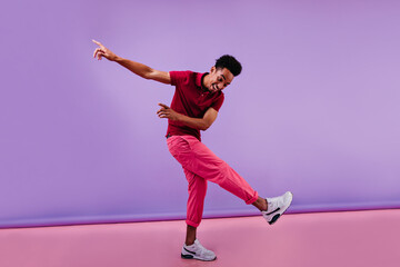 Laughing black guy with stylish haircut expressing emotions on purple background. Studio full-length portrait of african man dancing with sincere smile.