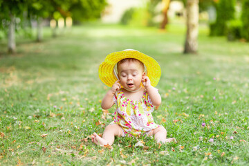 little baby girl 7 months old sitting on the green grass in a yellow dress and hat with her eyes closed and holding her hands to her ears, walking in the fresh air