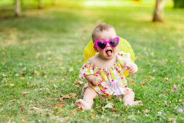 a small baby girl of 7 months is sitting on the green grass in a yellow dress, hat and bright glasses and showing her tongue, walking in the fresh air. Space for text