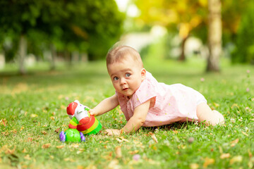 little baby girl 7 months old playing on a green lawn in a pink bodysuit, walking in the fresh air, early development of children up to a year