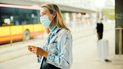 Young blonde woman wearing mask waiting in a bus station and using phone.