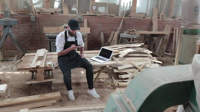 Carpenter in his own woodshop using a laptop or pc and writes notes while being  in his workspace. Small business concept.