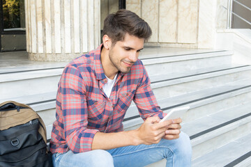 Young handsome student reading an electronic book with a cute smile while sitting on the stairs 